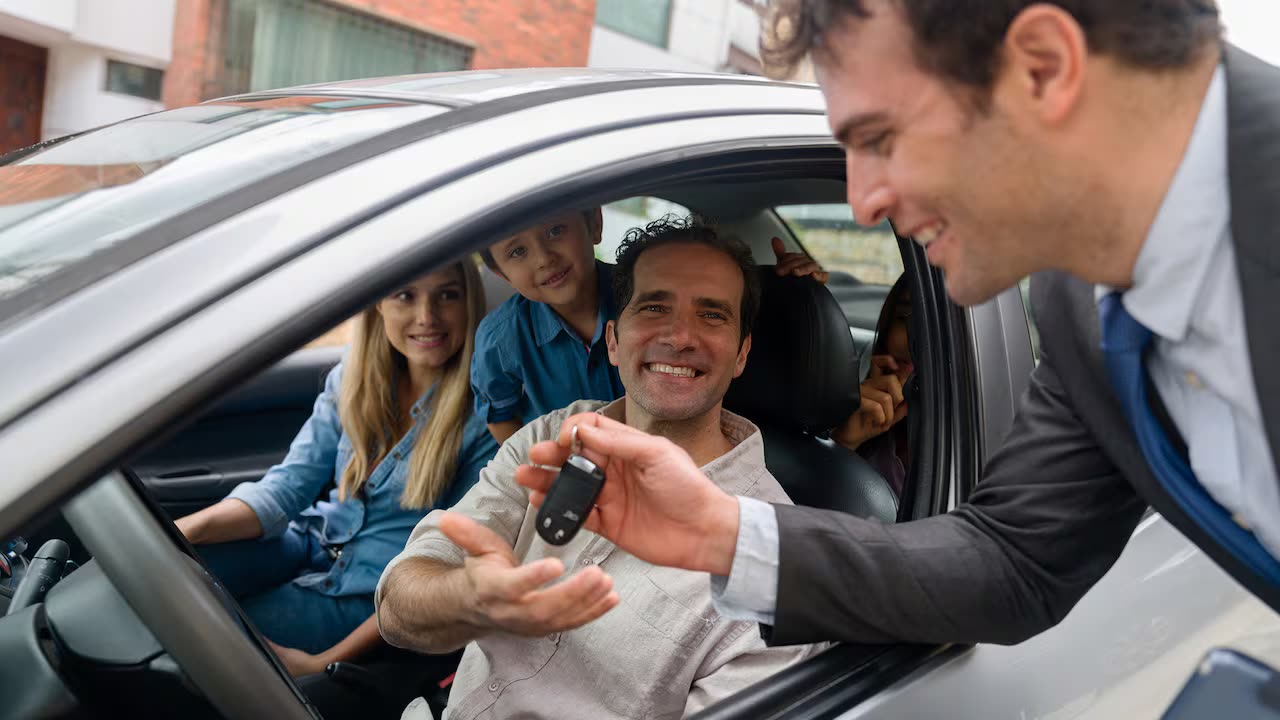 Un vendedor entrega la llave de un auto a una familia sentada dentro del vehículo